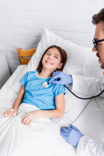 Pédiatre examen gai fille avec stéthoscope à l'hôpital, flou au premier plan — Photo de stock