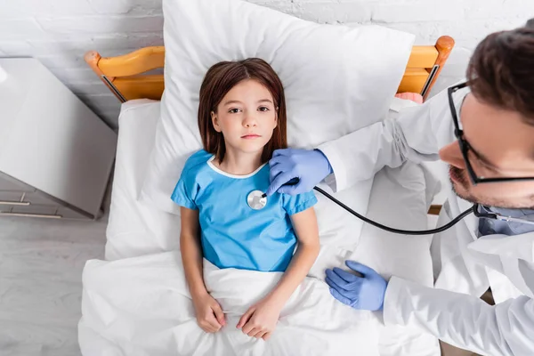 Над головой вид девушки лежащей в постели, когда врач осматривает ее стетоскопом — стоковое фото
