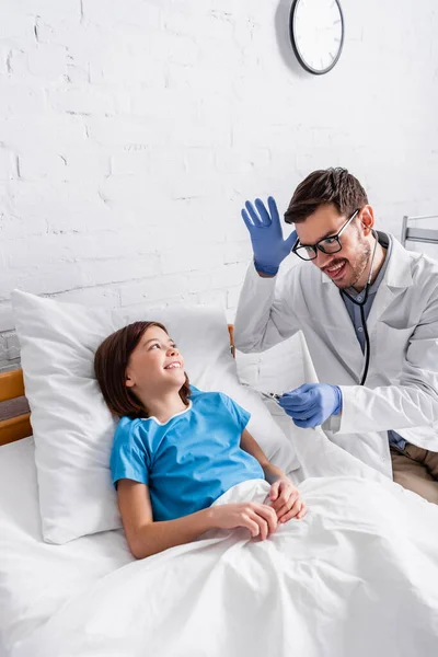 Alegre pediatra haciendo gestos y sacando la lengua mientras sostiene el termómetro cerca de la chica feliz acostada en la cama - foto de stock