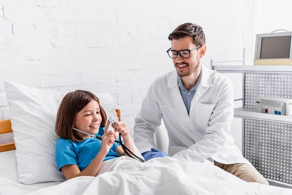 Niño feliz sosteniendo estetoscopio cerca de pediatra sonriente - foto de stock