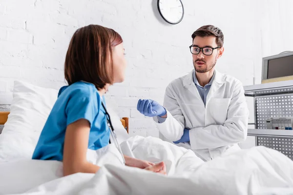 Pediatrician in latex gloves gesturing while talking to child on blurred foreground — Stock Photo