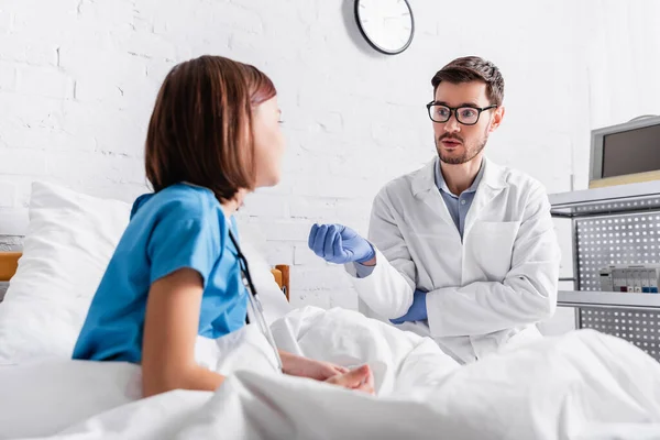 Pediatra en guantes de látex haciendo gestos mientras habla con el niño en primer plano borroso - foto de stock