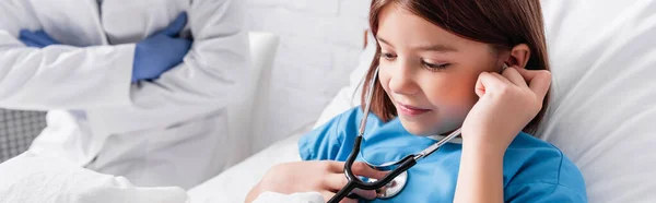 Smiling girl examining herself with stethoscope near doctor on blurred background, banner - foto de stock