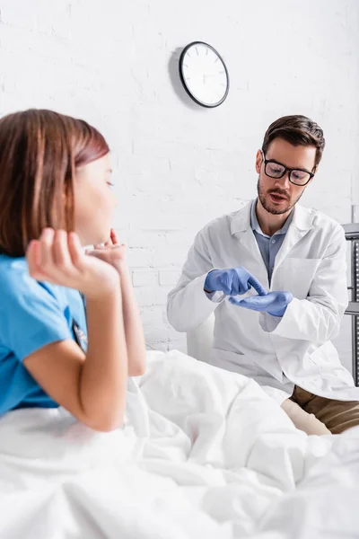 Médico apontando com o dedo na palma da mão enquanto conversa com a menina na cama em primeiro plano turvo — Fotografia de Stock