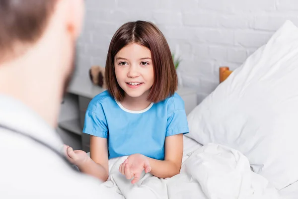 Lächelndes Mädchen gestikuliert im Gespräch mit Kinderarzt im verschwommenen Vordergrund — Stockfoto