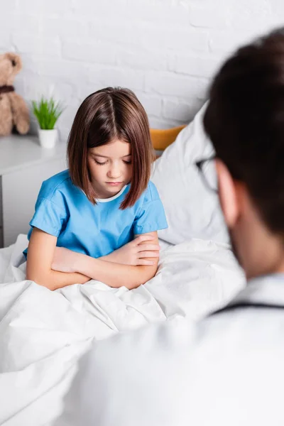 Chica molesta con los brazos cruzados en la cama del hospital cerca del médico en primer plano borrosa - foto de stock