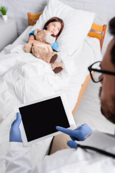 Foyer sélectif du médecin en utilisant une tablette numérique près d'une fille malade couchée au lit avec un ours en peluche — Photo de stock