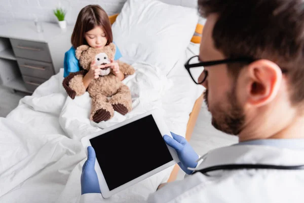 Foco seletivo de tablet digital com tela em branco nas mãos do pediatra perto da menina segurando ursinho na cama no hospital — Fotografia de Stock