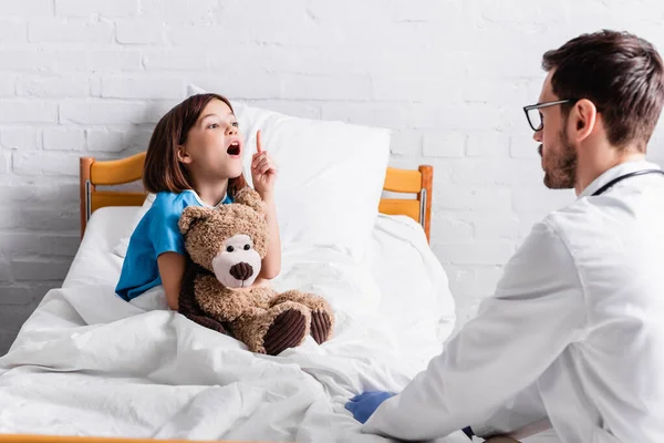Aufgeregtes Mädchen hält Teddybär in der Hand und zeigt Ideengeste beim Kinderarzt — Stockfoto