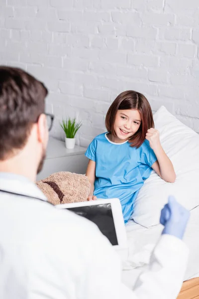 Glückliches Kind zeigt Gewinngeste beim Kinderarzt mit digitalem Tablet im verschwommenen Vordergrund — Stockfoto