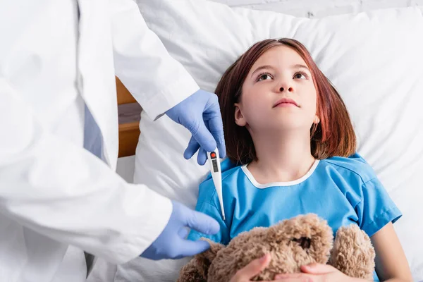 Medico in guanti di lattice che tiene il termometro vicino al bambino malato sdraiato con giocattolo a letto — Foto stock