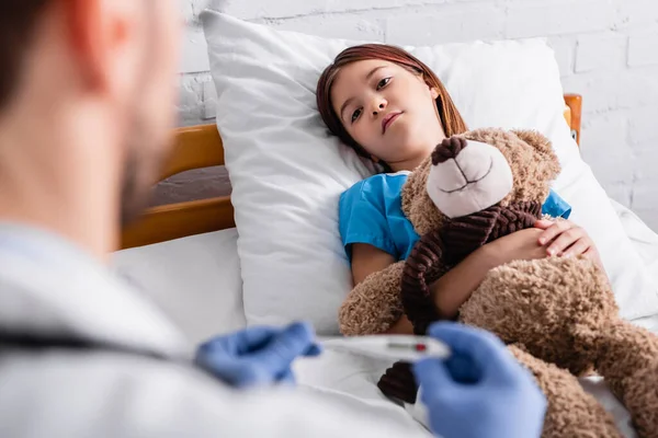 Criança doente deitada na cama com ursinho perto do pediatra com termômetro em primeiro plano borrado — Fotografia de Stock