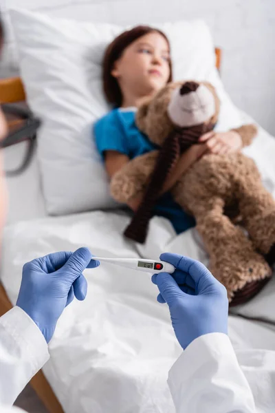 Foco selectivo del termómetro en manos del pediatra cerca del niño enfermo acostado en la cama con el oso de peluche - foto de stock