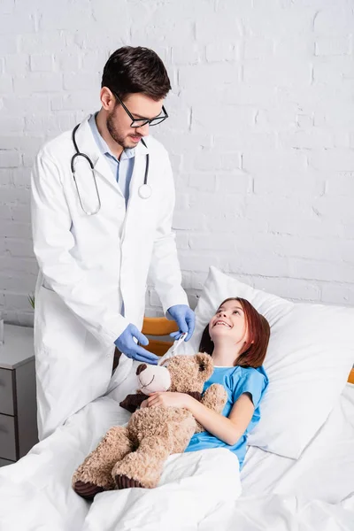 Pediatra segurando termômetro perto menina feliz deitado na cama com ursinho de pelúcia — Fotografia de Stock