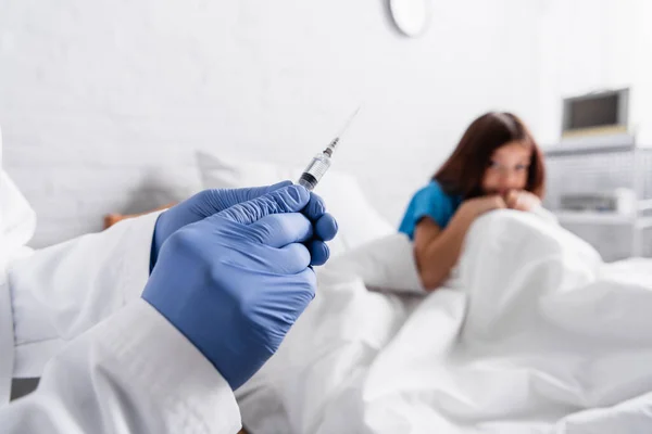 Afraid child sitting on bed near doctor with syringe on blurred foreground — Stock Photo
