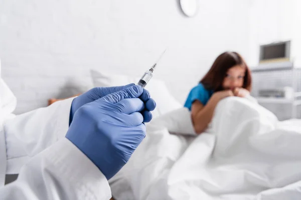 Niño asustado sentado en la cama cerca del médico con jeringa en primer plano borroso - foto de stock