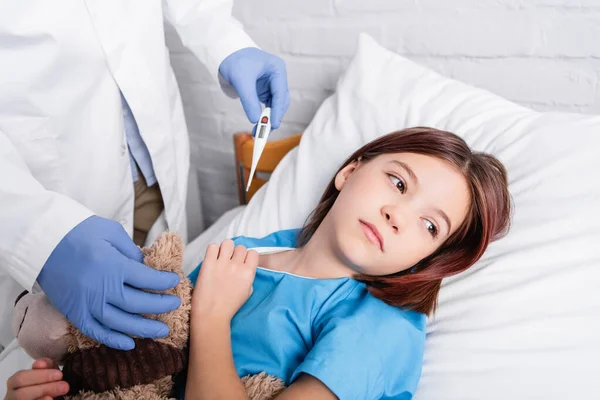 Doctor en guantes de látex sosteniendo termómetro cerca de niño acostado en la cama con osito de peluche - foto de stock