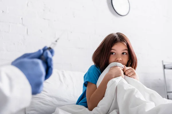 Arzt hält Spritze neben verängstigtem Mädchen, das unter Decke auf Bett sitzt — Stockfoto