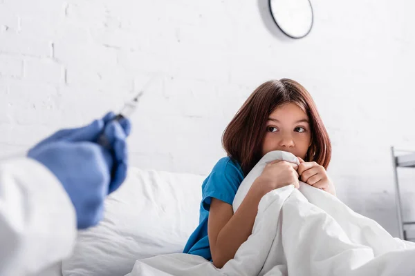 Médico sosteniendo la jeringa cerca de chica asustada sentada en la cama debajo de la manta - foto de stock