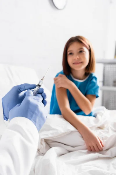 Pediatrician holding syringe near smiling child sitting on bed — Stock Photo