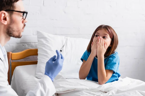 Chica asustada cubriendo la boca con las manos cerca de pediatra con jeringa en primer plano borrosa - foto de stock