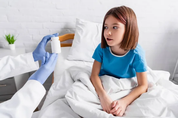 Médico segurando recipiente com medicamentos perto menina espantada na cama do hospital — Fotografia de Stock