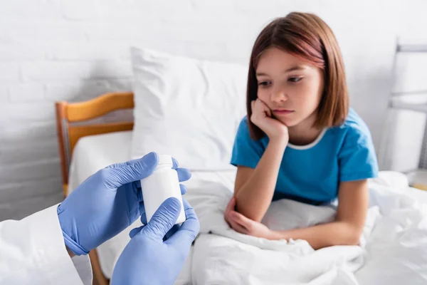 Verärgertes krankes Mädchen im Krankenhausbett in der Nähe von Arzt, der Behälter mit Medikamenten hält — Stockfoto