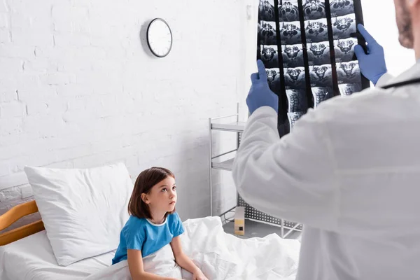 Médecin tenant une radiographie près d'un enfant assis au lit à l'hôpital, avant-plan flou — Photo de stock