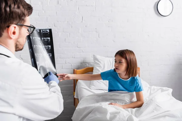 Chica sentada en la cama y apuntando a la radiografía en manos del médico en primer plano borrosa - foto de stock