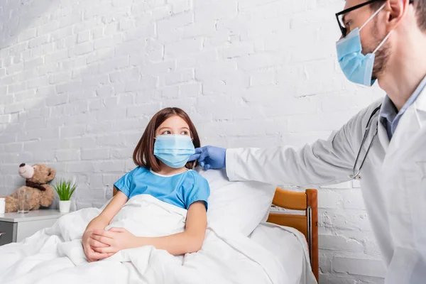 Kinderarzt in medizinischer Maske berührt Hals eines kranken Mädchens im Krankenhausbett — Stockfoto