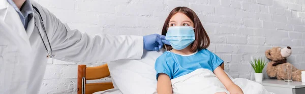 Врач трогает шею ребенка лежащего в постели в медицинской маске, баннер — стоковое фото