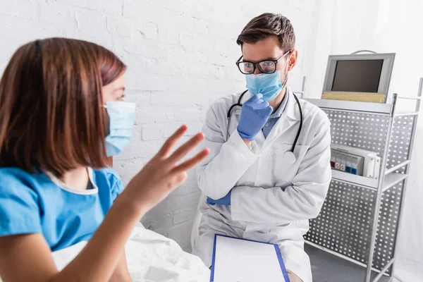 Menina doente em máscara médica mostrando três dedos enquanto conversa com pediatra no hospital — Fotografia de Stock