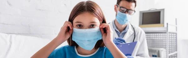 Sick kid fixing medical mask while looking at camera near doctor writing diagnosis on blurred background, banner - foto de stock