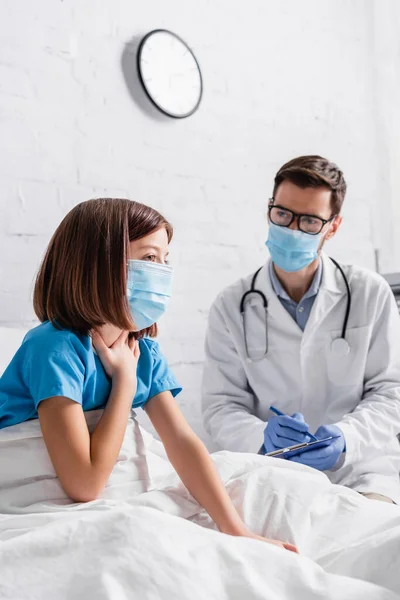 Girl in medical mask touching throat near doctor writing diagnosis on clipboard — Stock Photo