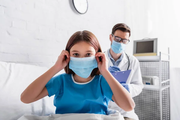 Krankes Mädchen blickt in Kamera, während es medizinische Maske in der Nähe des Arztes fixiert, der Rezept auf verschwommenem Hintergrund schreibt — Stockfoto