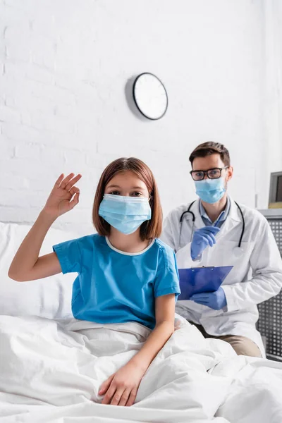 Criança em máscara médica mostrando gesto ok perto do médico com área de transferência em fundo borrado — Fotografia de Stock