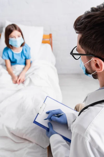 Kinderarzt schreibt Diagnose auf Klemmbrett neben krankem Mädchen in medizinischer Maske auf verschwommenem Hintergrund — Stockfoto