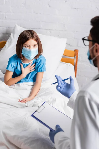 Child in medical mask touching chest while talking to doctor with clipboard on blurred foreground — Stock Photo