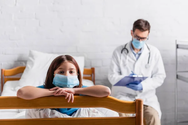 Sick girl in medical mask looking at camera near doctor writing on clipboard on blurred background — Stock Photo