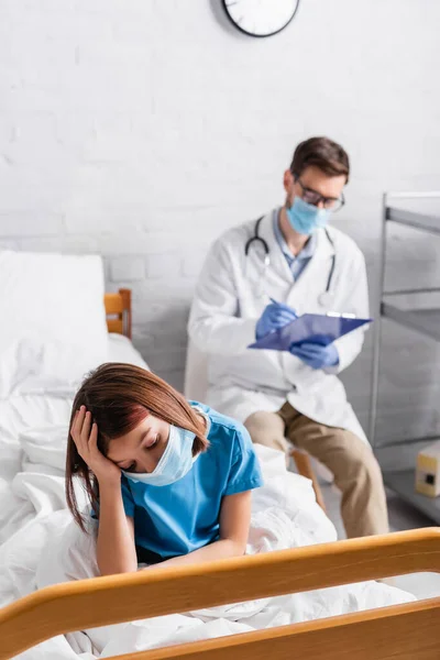 Больной ребенок в медицинской маске страдает от головной боли в то время как доктор пишет диагноз на размытом фоне — стоковое фото