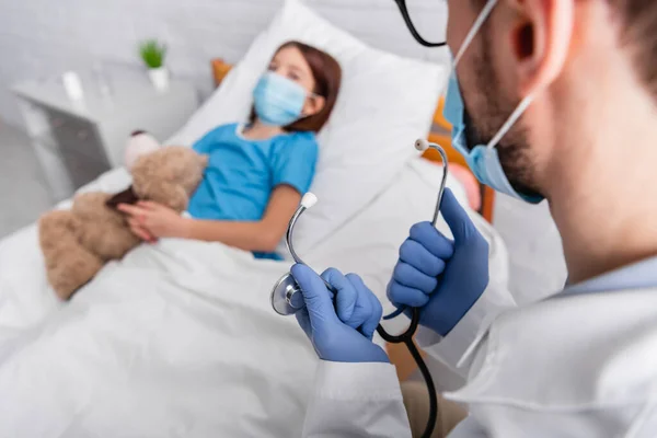 Médico segurando estetoscópio perto de criança doente em máscara médica deitada na cama com ursinho de pelúcia no fundo borrado — Fotografia de Stock