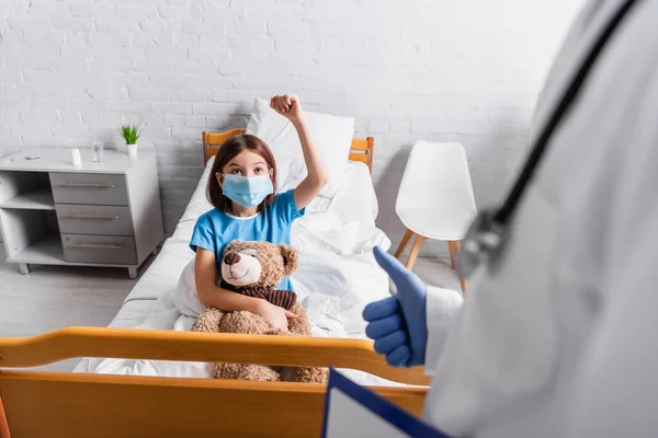 Krankes Mädchen zeigt Siegergeste in der Nähe des Arztes mit erhobenem Daumen im verschwommenen Vordergrund — Stockfoto