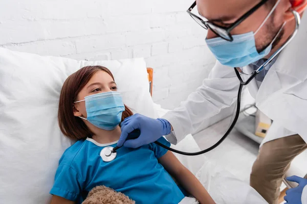 Pediatra en máscara médica examinando niña enferma con estetoscopio en el hospital - foto de stock