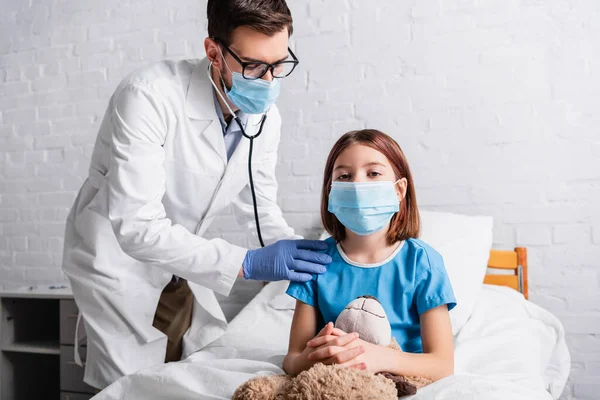 Chica enferma en máscara médica con osito de peluche cerca pediatra examinándola con estetoscopio - foto de stock