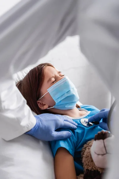 Fille malade dans le masque médical couché avec les yeux fermés près du médecin l'examinant avec stéthoscope, avant-plan flou — Photo de stock