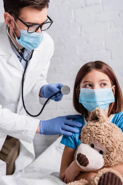 Pediatra com estetoscópio perto da menina em máscara médica sentado na cama com ursinho de pelúcia — Fotografia de Stock