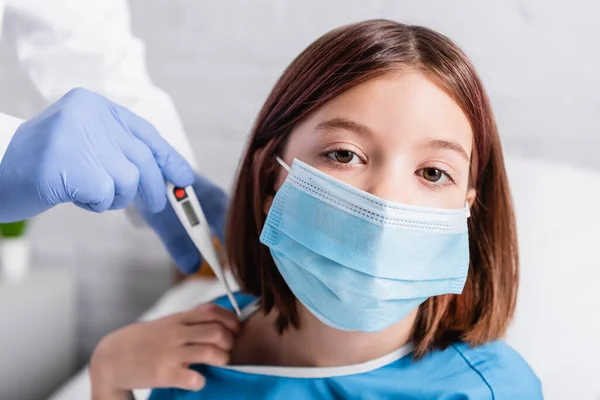 Chica enferma en máscara médica mirando a la cámara cerca del médico con termómetro sobre fondo borroso - foto de stock