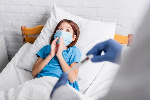 Shocked girl in medical mask showing pray gesture near pediatrician with thermometer on blurred foreground — Stock Photo