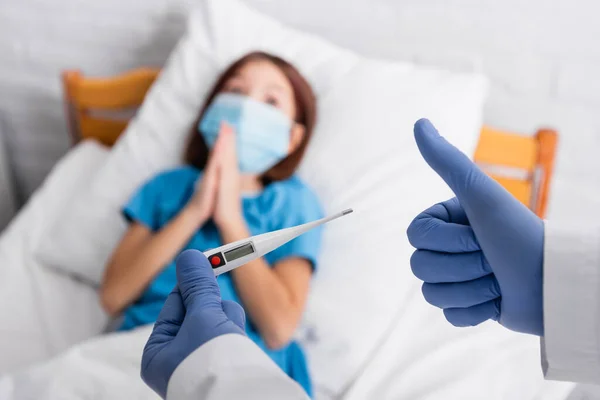 Doctor holding thermometer and showing thumb up near child in medical mask lying in bed with praying hands, blurred background - foto de stock