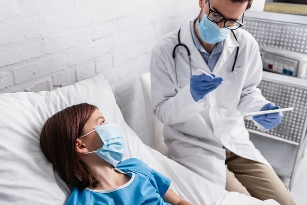 Лікар в медичній масці, дивлячись на термометр, тримаючи цифровий планшет біля хворої дитини — стокове фото